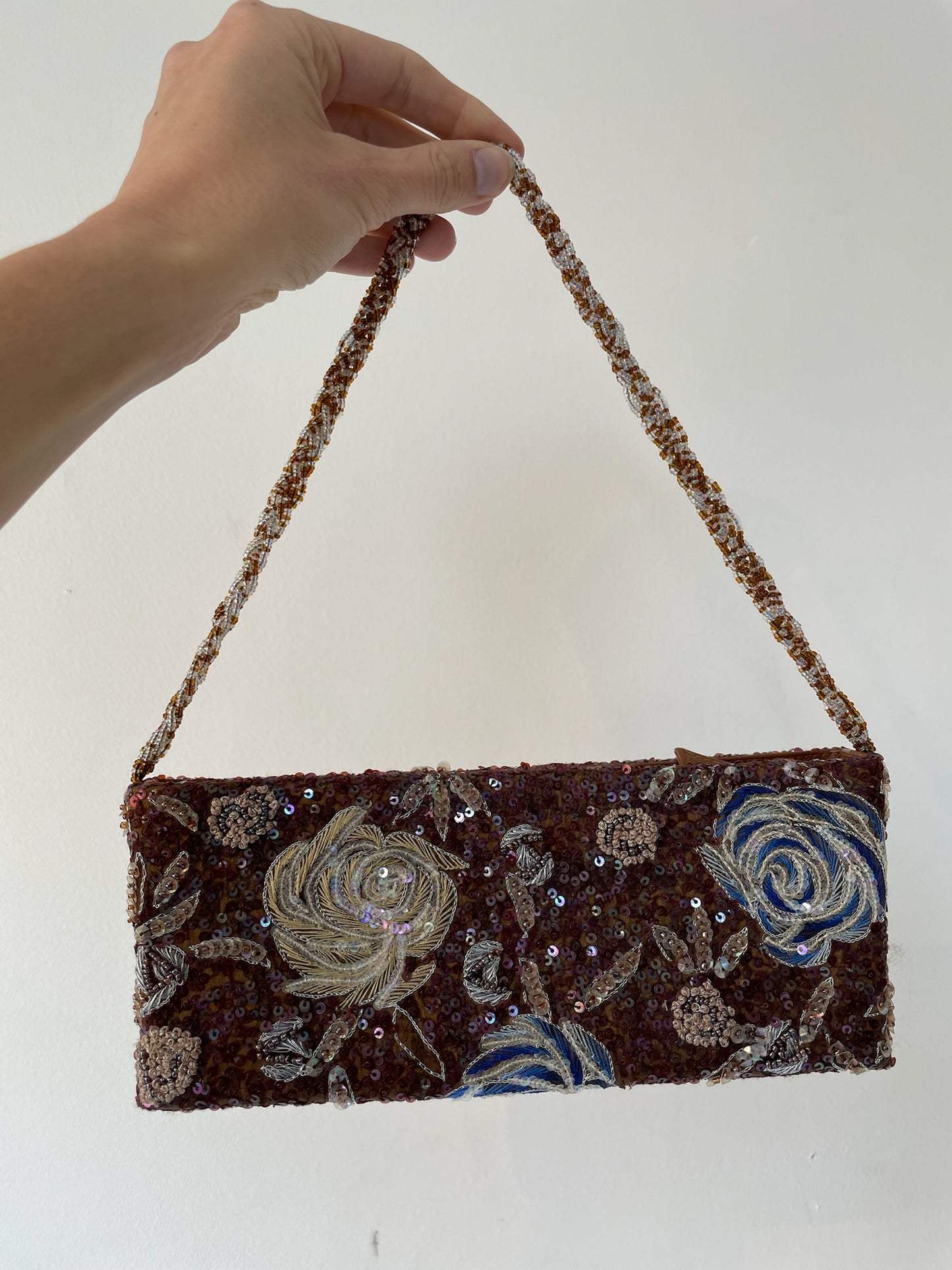 Hand holding a brown handbag with floral embroidery and sequins on a white background
