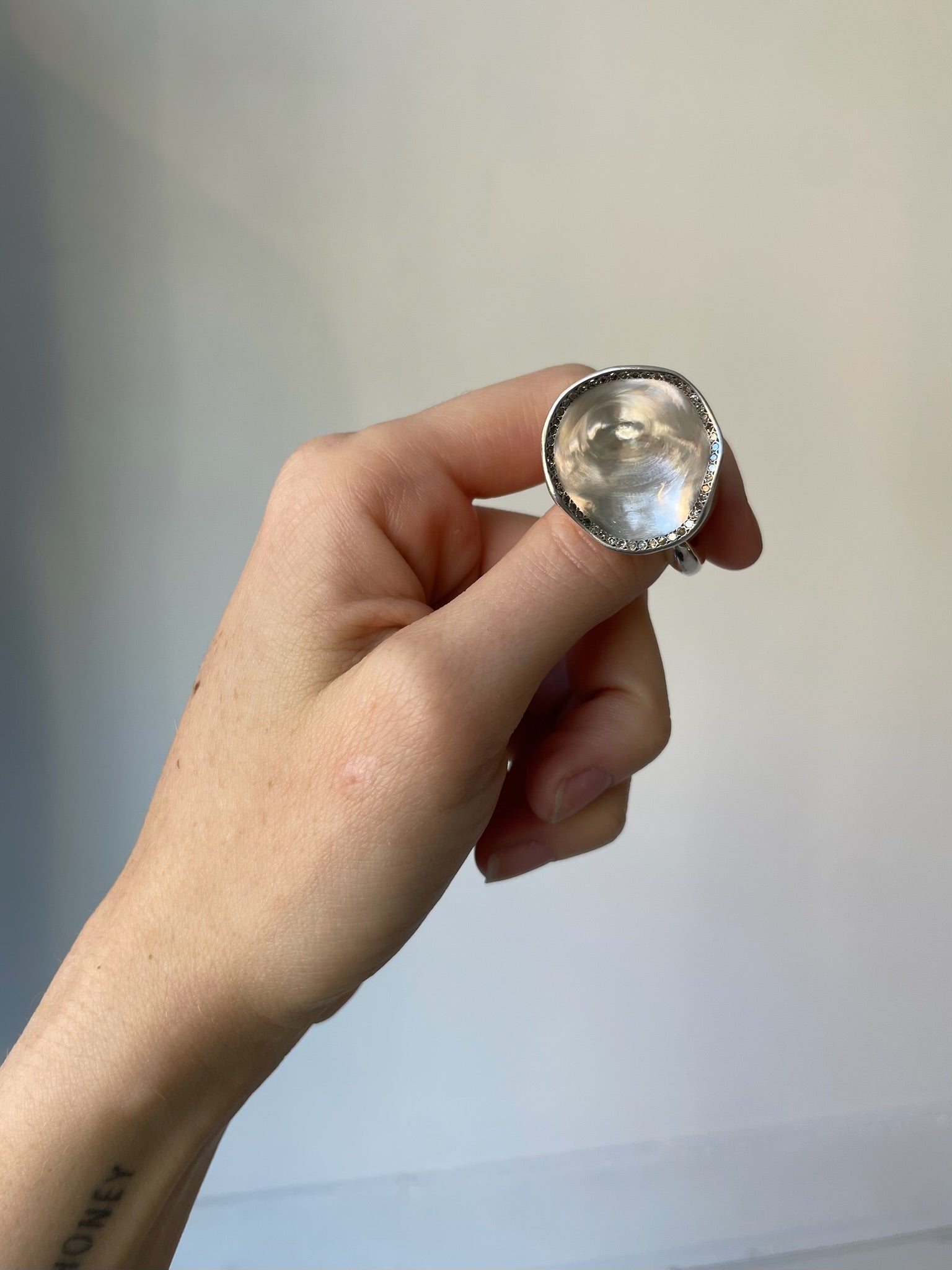 Hand holding a silver ring with a large gemstone against a plain background